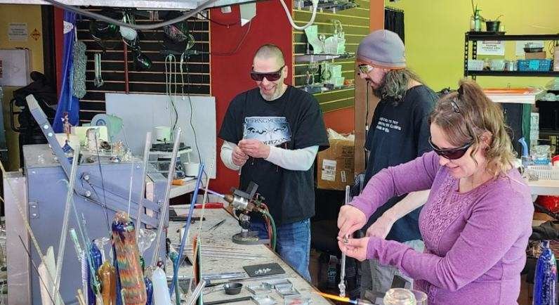 Three people working together at a glass blowing workbench